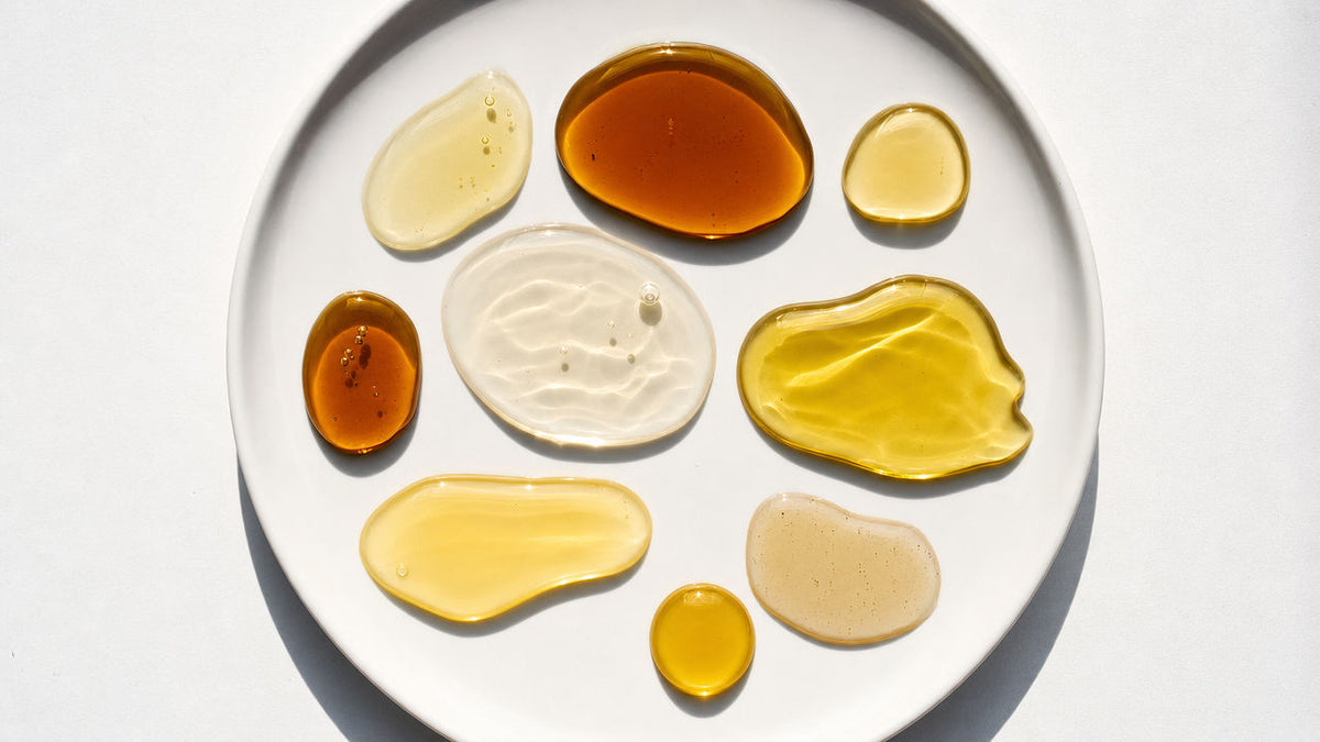 Comparison of different hair oils for growth showing coconut oil, argan oil, castor oil, jojoba oil, and rosemary oil pooled on a white ceramic plate with varying viscosities and amber golden colors