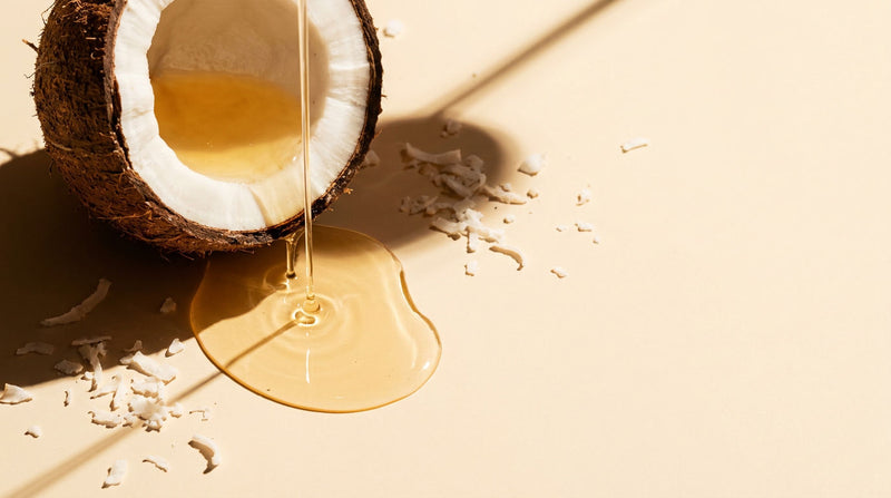 Halved fresh coconut with golden coconut oil streaming and pooling on a warm cream surface, showing the clear amber oil used for hair growth and protein loss prevention
